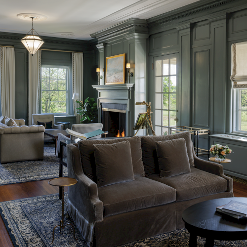 A stylish living room with gray sofas, rugs, lamps, and a lit fireplace, framed by large windows and classic molding.