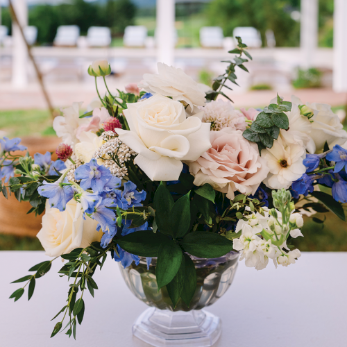 A glass vase holds a mixed bouquet of white roses, blue flowers, and greenery at an outdoor wedding venue.