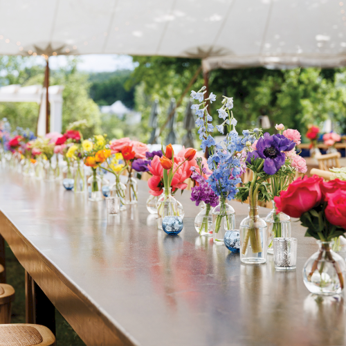 A long outdoor event table lined with colorful bouquets in glass vases under a white canopy, with chairs all around.