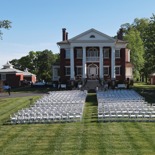 An outdoor ceremony setup with white chairs facing a grand brick mansion on a sunny day, with trees and tents nearby.