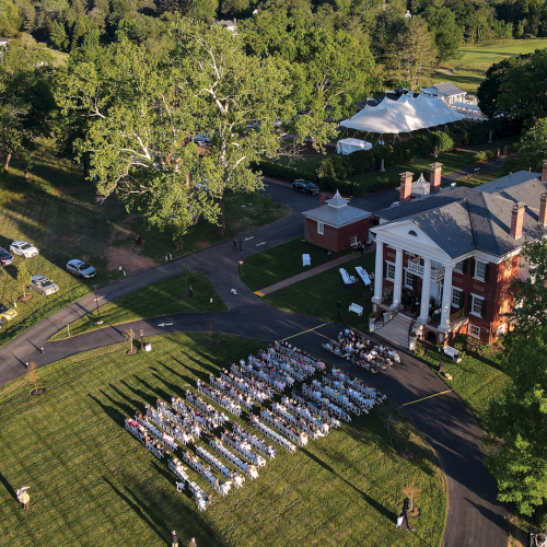 Aerial view of a countryside estate with a big tent, wedding guests seated in rows of chairs, and parked cars by the mansion.