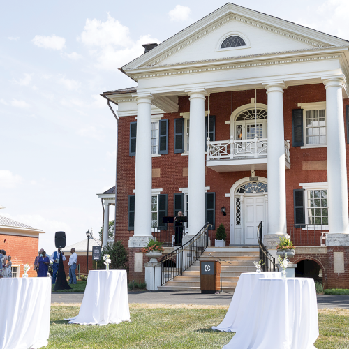A brick colonial mansion with tall white columns hosts an outdoor gathering with white-covered tables on a sunny lawn.