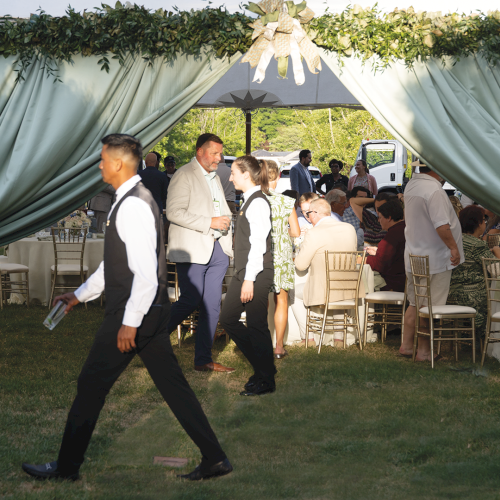 An outdoor catered event under a decorated canopy tent, with guests mingling around gold chairs and a table setup.