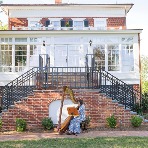 A woman plays a harp on the steps of a red-brick building while trees fill the sunny courtyard.