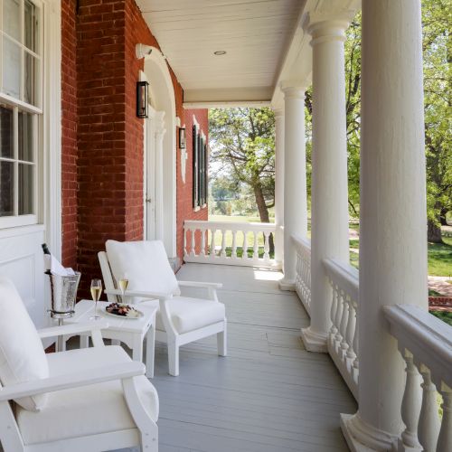 A sunny front porch with white rocking chairs, a small table set with drinks and fruit, and red brick pillars overlooking greenery.