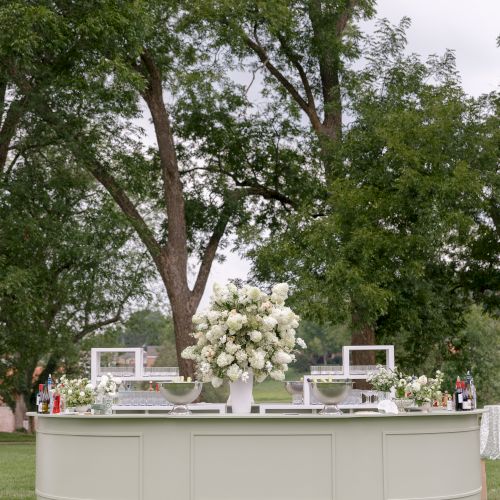 An outdoor event bar with clear glasses, bottles, and white floral centerpieces in a lush green park.