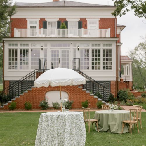 Outdoor wedding-style setup with decorated tables and chairs, a white umbrella, and a brick building in the background.