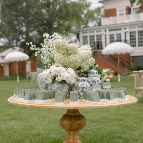 An outdoor wedding escort table with white flowers, ornate vases, place cards, and umbrellas beside a brick building.