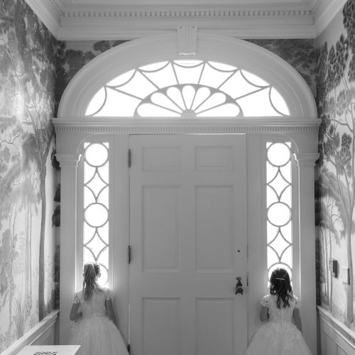 Black-and-white interior hallway with a large paneled door and arched stained-glass window, flanked by two girls in dresses.