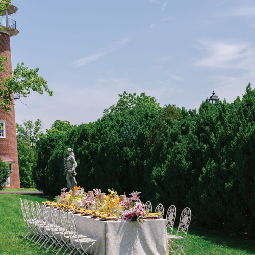 Outdoor garden scene with a brick tower, statues, manicured hedges, and a banquet table set with flowers and plates.