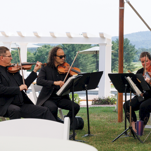 An outdoor string quartet plays violins and cello on a garden stage, surrounded by umbrellas and scenic hills.