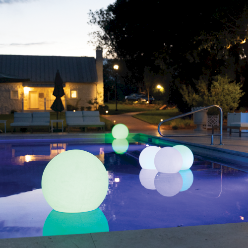 A nighttime backyard pool lit by glowing sphere lights, lounge chairs, and warm building lights under a dusky sky.