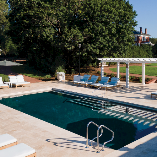 An outdoor pool with lounge chairs and white pergola, surrounded by green trees and a large house on a sunny day.
