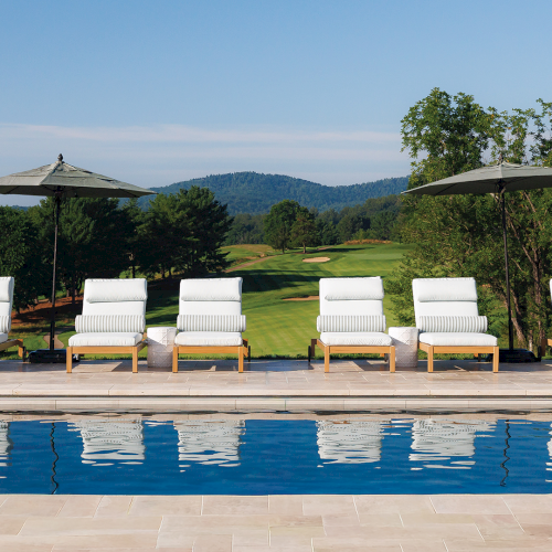 An outdoor pool with white lounge chairs and patio umbrellas overlooks rolling green hills and trees under a clear blue sky.