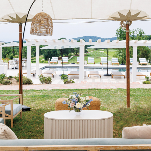 A sunny outdoor poolside patio with white pergola, hanging lamps, lounge chairs, a flowered table, and a grassy garden view.