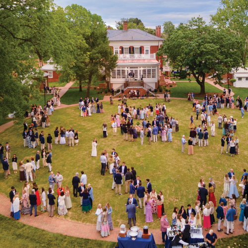 A large outdoor wedding reception with guests mingling on a grassy lawn in front of a grand brick mansion.