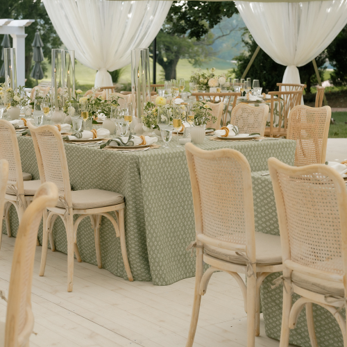 Outdoor wedding reception with long table settings, white draped canopies, floral centerpieces, candles, and wicker chairs.