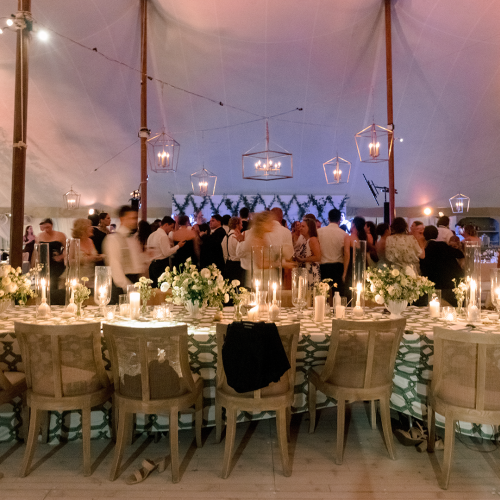 A decorated outdoor wedding reception with long tables of flowers and candles, surrounded by guests under glowing lanterns.