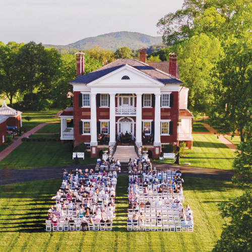 An aerial view shows a large historic mansion on green grounds with an outdoor ceremony setup and rows of chairs.