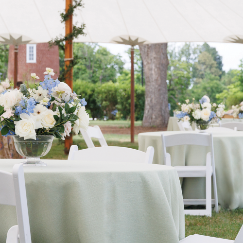 An outdoor wedding reception set-up with white chairs, round tables draped in fabric, and a bouquet centerpiece of blue and white flowers.
