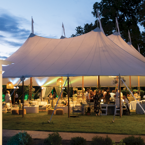 An outdoor event under a large white tent marquee at dusk, with lights, tables, and guests gathered inside.