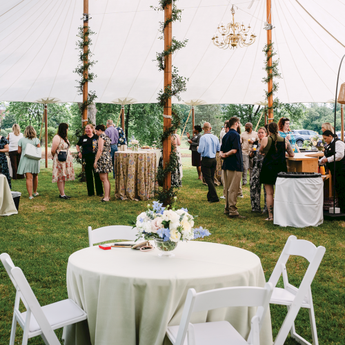 An outdoor garden party under a white tent, with guests mingling, floral centerpieces, and a decorated bar and chairs.