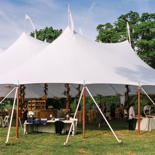 Outdoor event under large white pole tents with tables, chairs, and people gathering in a sunny garden.