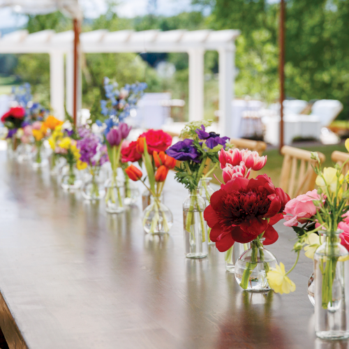 Colorful bouquets in glass vases line a long outdoor event table, with wooden chairs and greenery in the background.