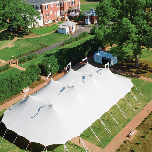 Aerial view of a large white event tent on a green campus lawn surrounded by trees and brick buildings.