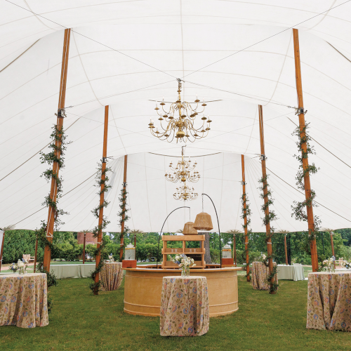 Outdoor event under a white marquee tent with hanging chandeliers, floral decor, and tables with white chairs.