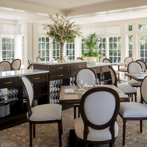 Elegant dining hall with set tables, glassware, flowers, and a waiter walking past.