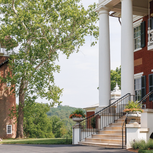 People stroll by a brick building with tall white columns and verandas, under lush trees beside a circular brick tower.