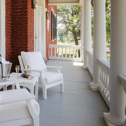 A sunny front porch with white rocking chairs, a small table set with drinks and snacks, and brick-and-column surroundings.