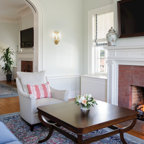 A bright living room with a wood-burning fireplace, chandelier lights, framed art, and upholstered chairs.