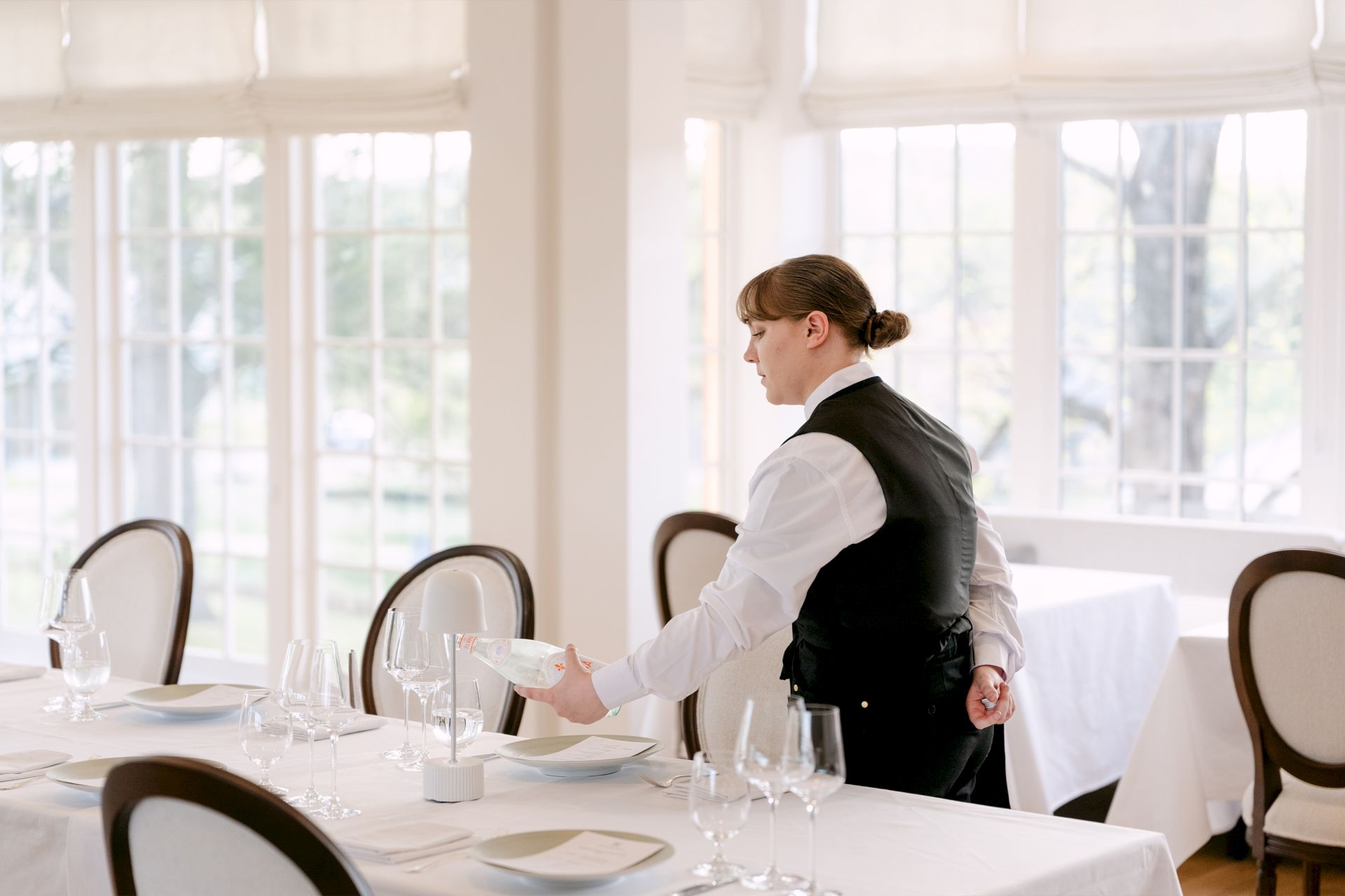 A waiter in a bright dining room pours a drink for guests at a table set with glassware and cutlery.