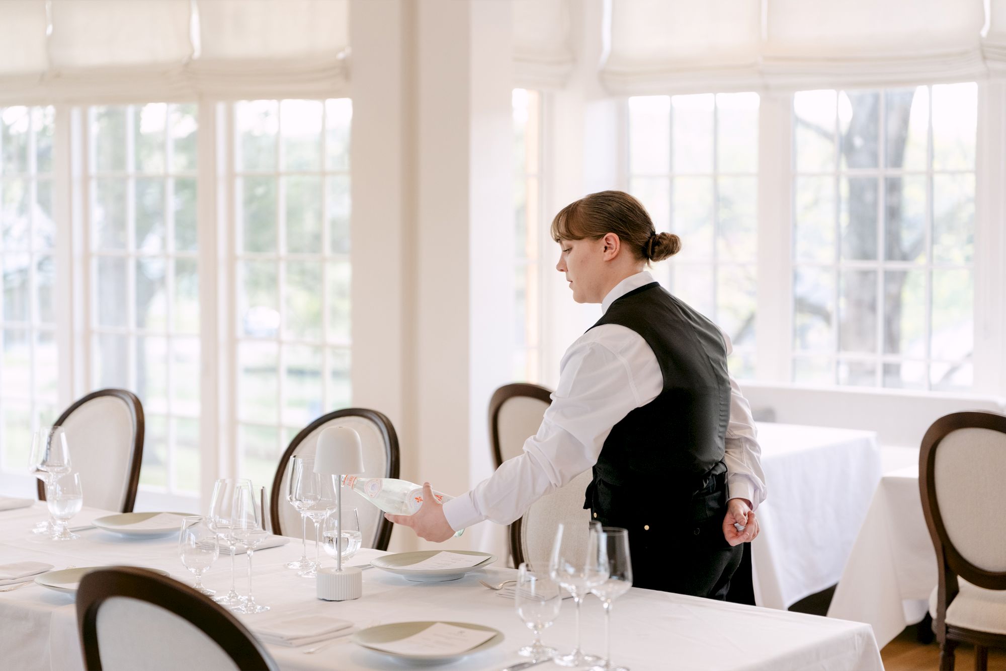 A waiter in a bright dining room pours a drink for guests at a table set with glassware and cutlery.