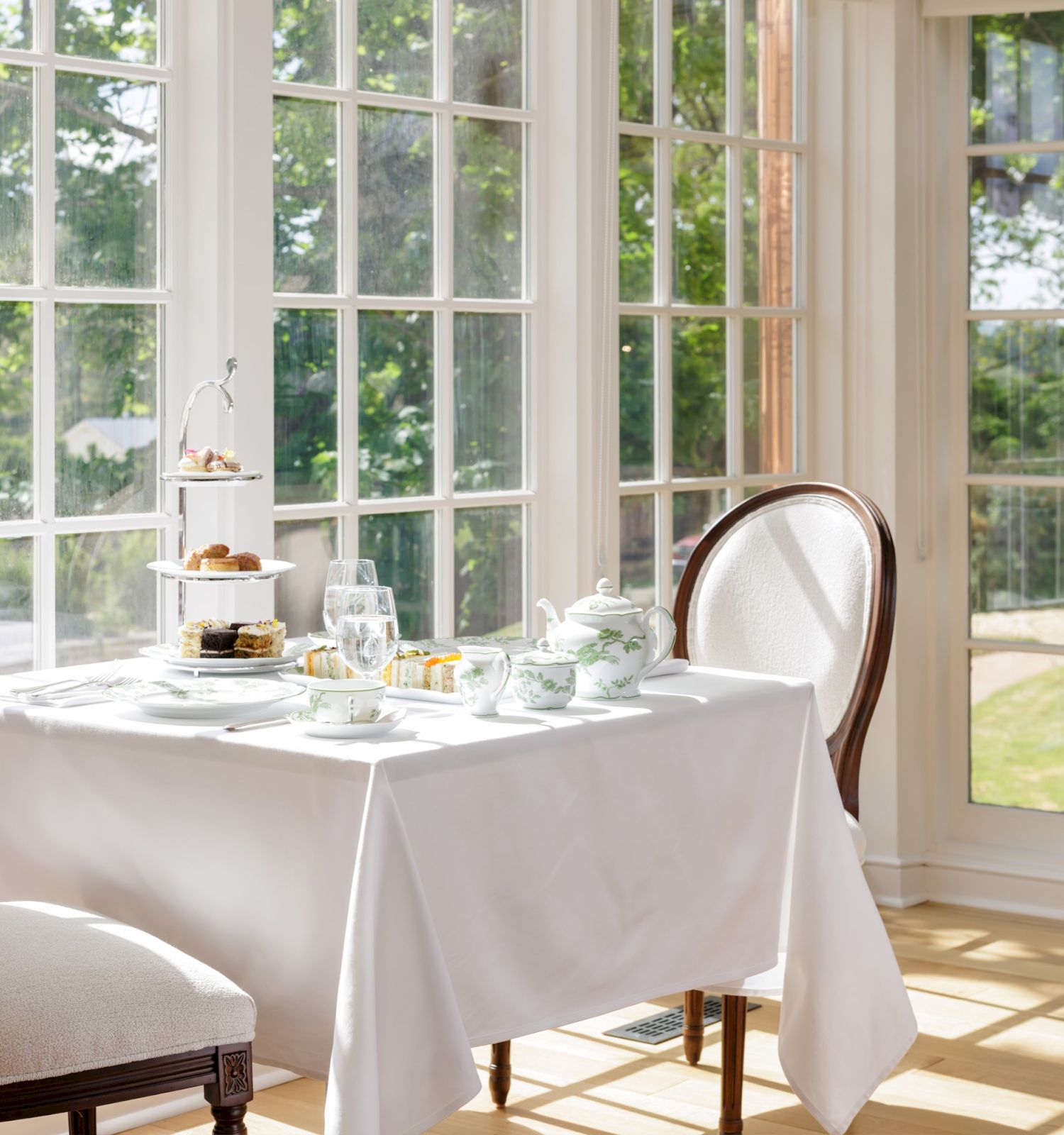 A bright window-side afternoon tea setup: white tablecloth, tiered cakes, teapot, cups, and a glass by potted plants.