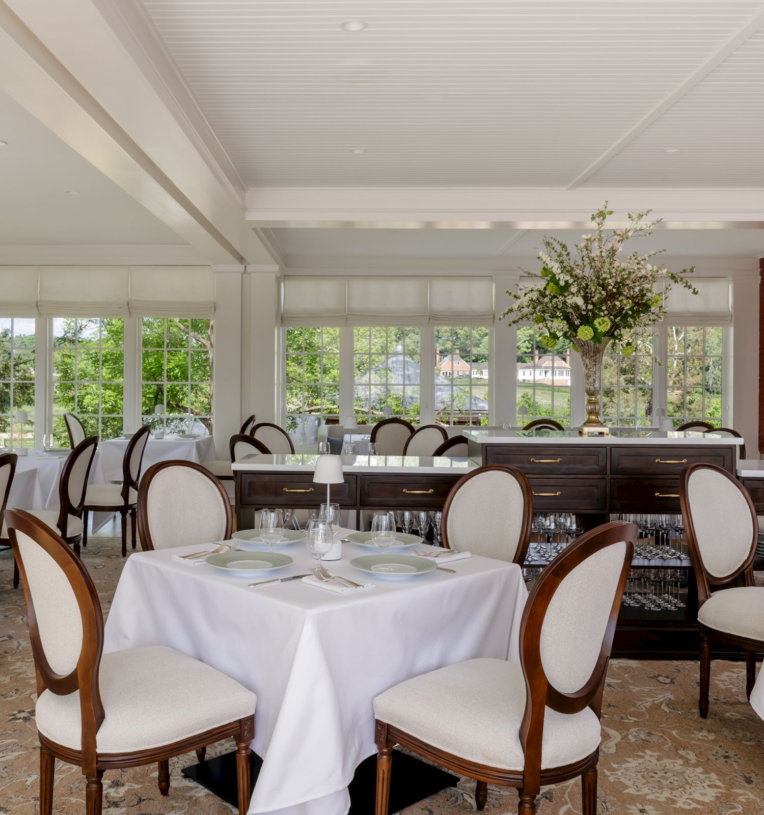 A bright banquet dining room with neatly set tables, chairs, glassware, and a large flower centerpiece.