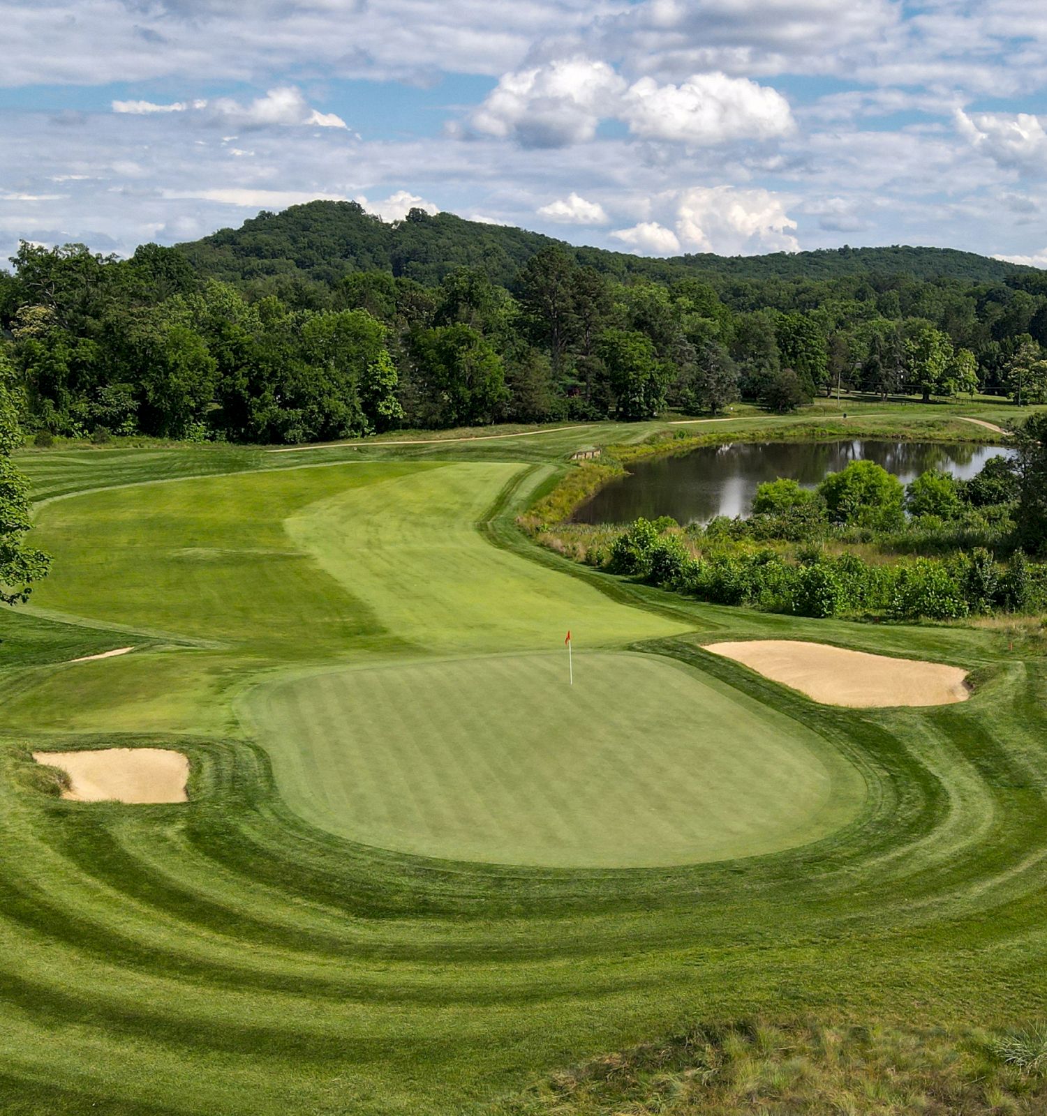 A scenic golf course fairway with sand bunkers and a pond, surrounded by lush green trees and rolling hills under a cloudy sky.