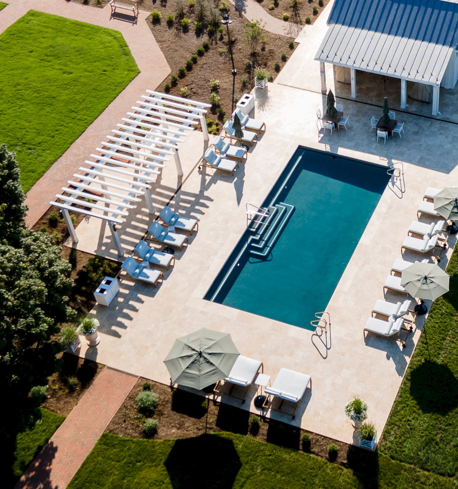 Aerial view of a sunny resort courtyard with a blue swimming pool, sun loungers, umbrellas, and manicured green gardens.