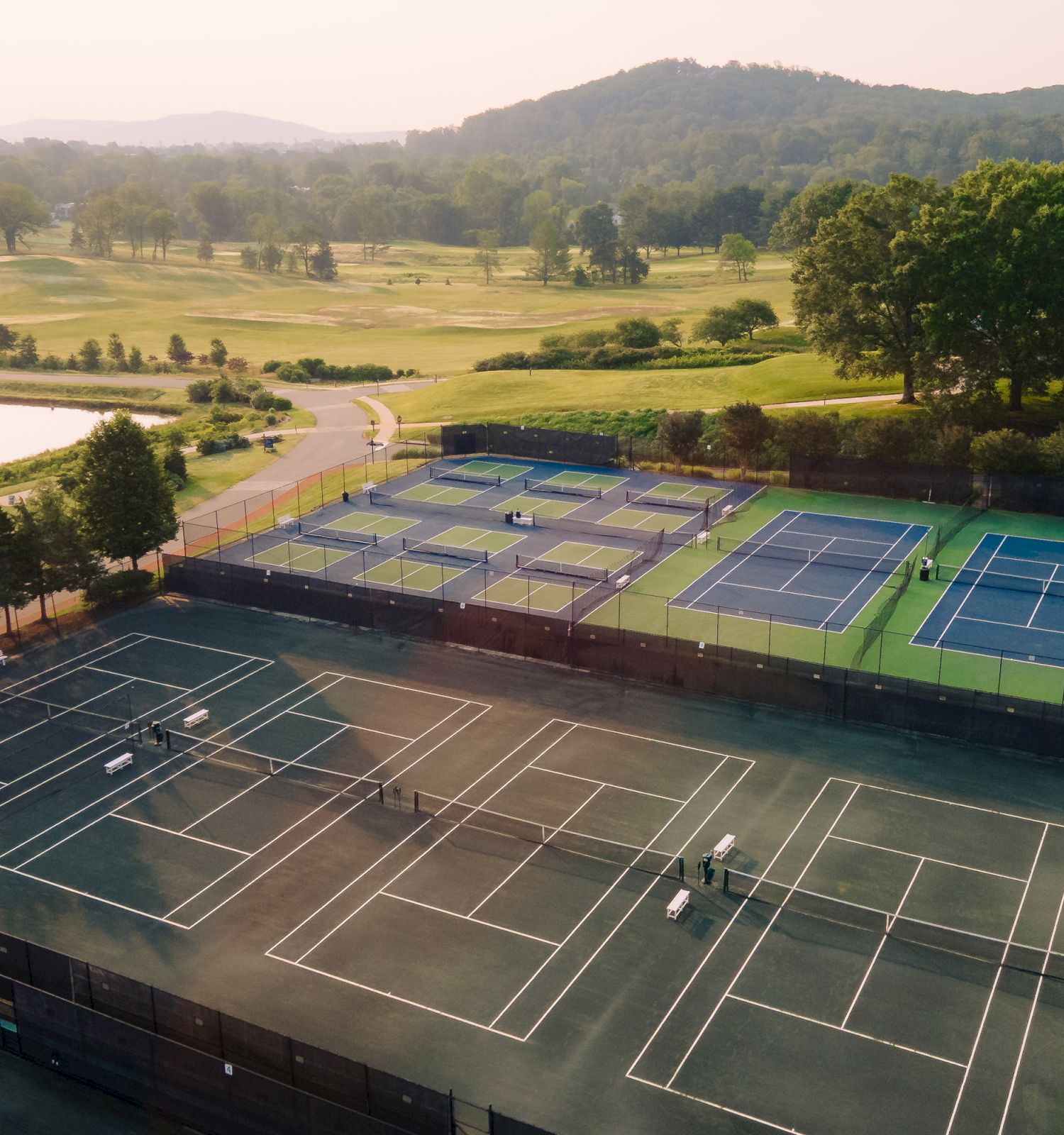 Aerial view of multiple tennis courts and a paddle-court complex beside a pond, surrounded by green fields and trees at dusk.