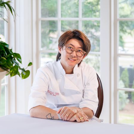 A woman chef in a white coat with glasses sits at a table by a window with greenery, smiling.