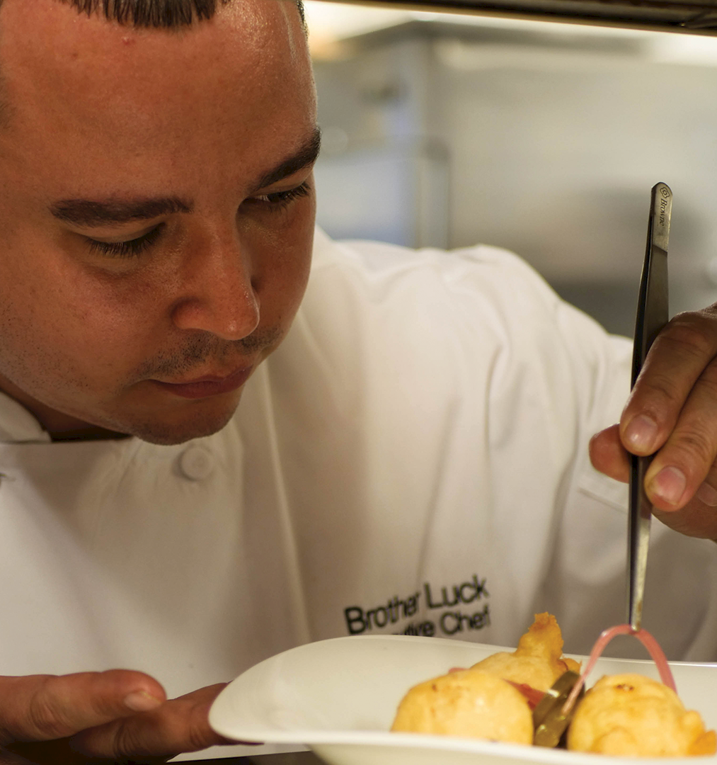 A chef in a white coat tastes food on a plate, holding a spoon and fork in a restaurant kitchen.