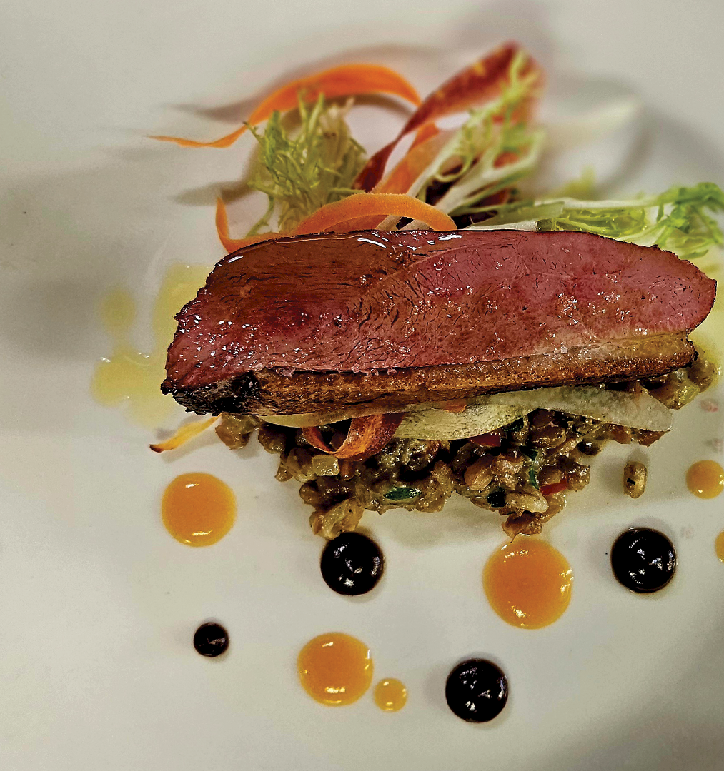 A plated seared beef steak with greens and crispy garnish, surrounded by dots of sauces on a white dish.