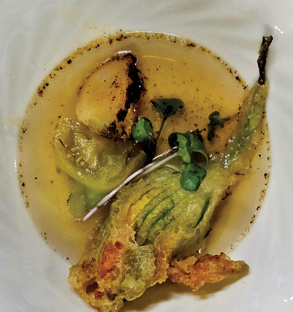 A white bowl of yellow broth with charred meat and green herb garnish, on the table.