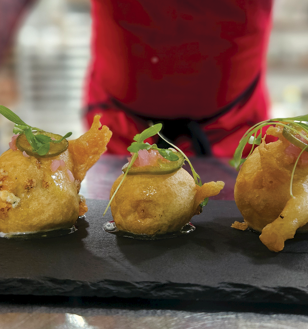 Three golden fried dumplings with herb garnish sit on a slate plate as a person in red stands behind them.