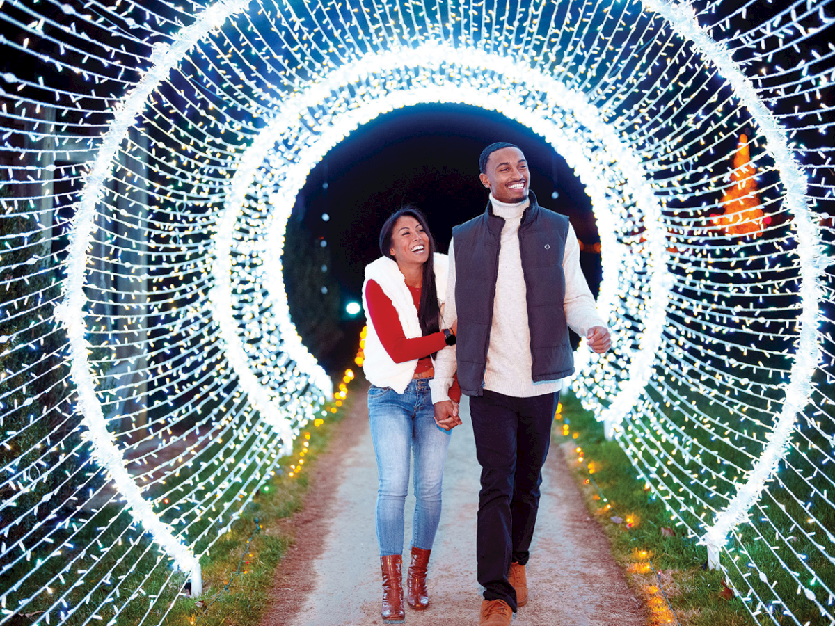 A smiling couple walks hand in hand through a glowing tunnel of white string lights.