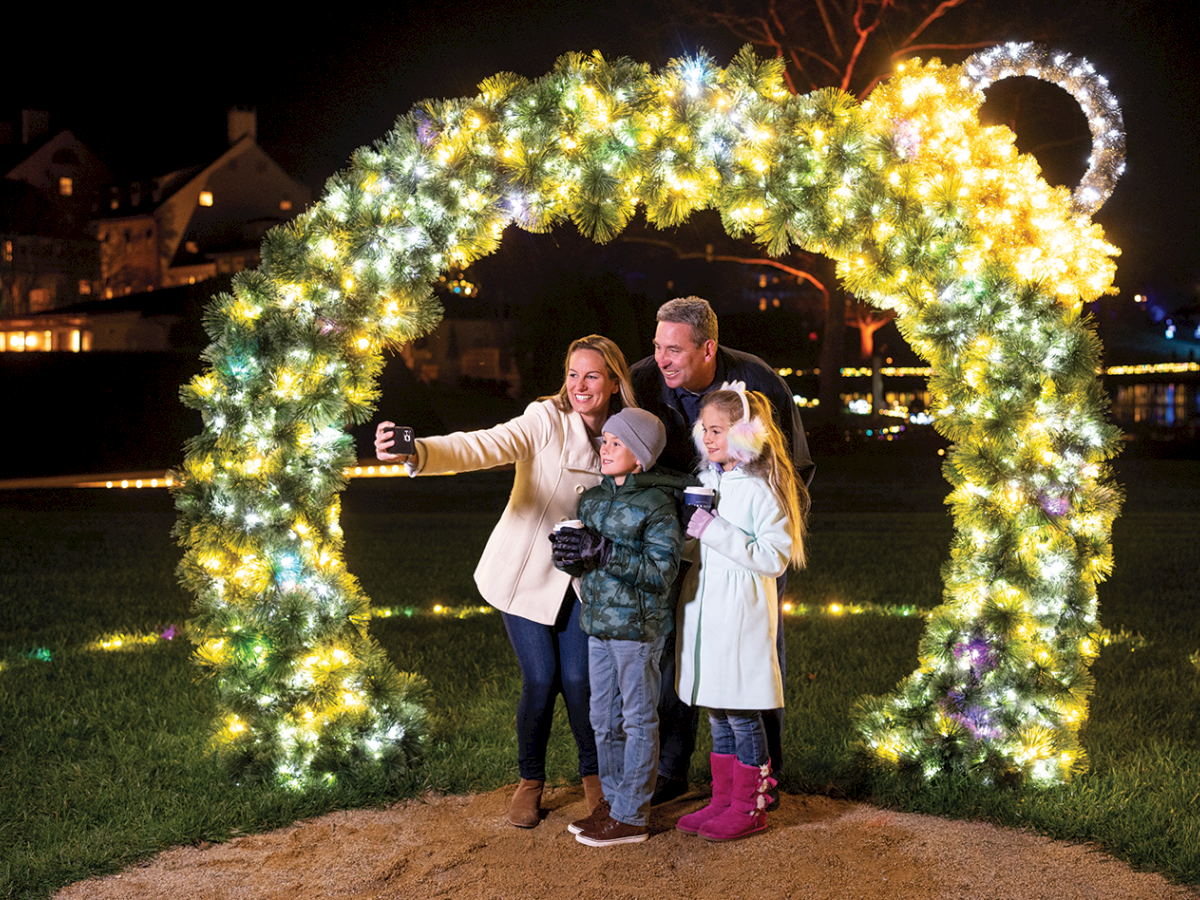 A family poses for a selfie under a glowing Christmas wreath of lights at night, smiling together.