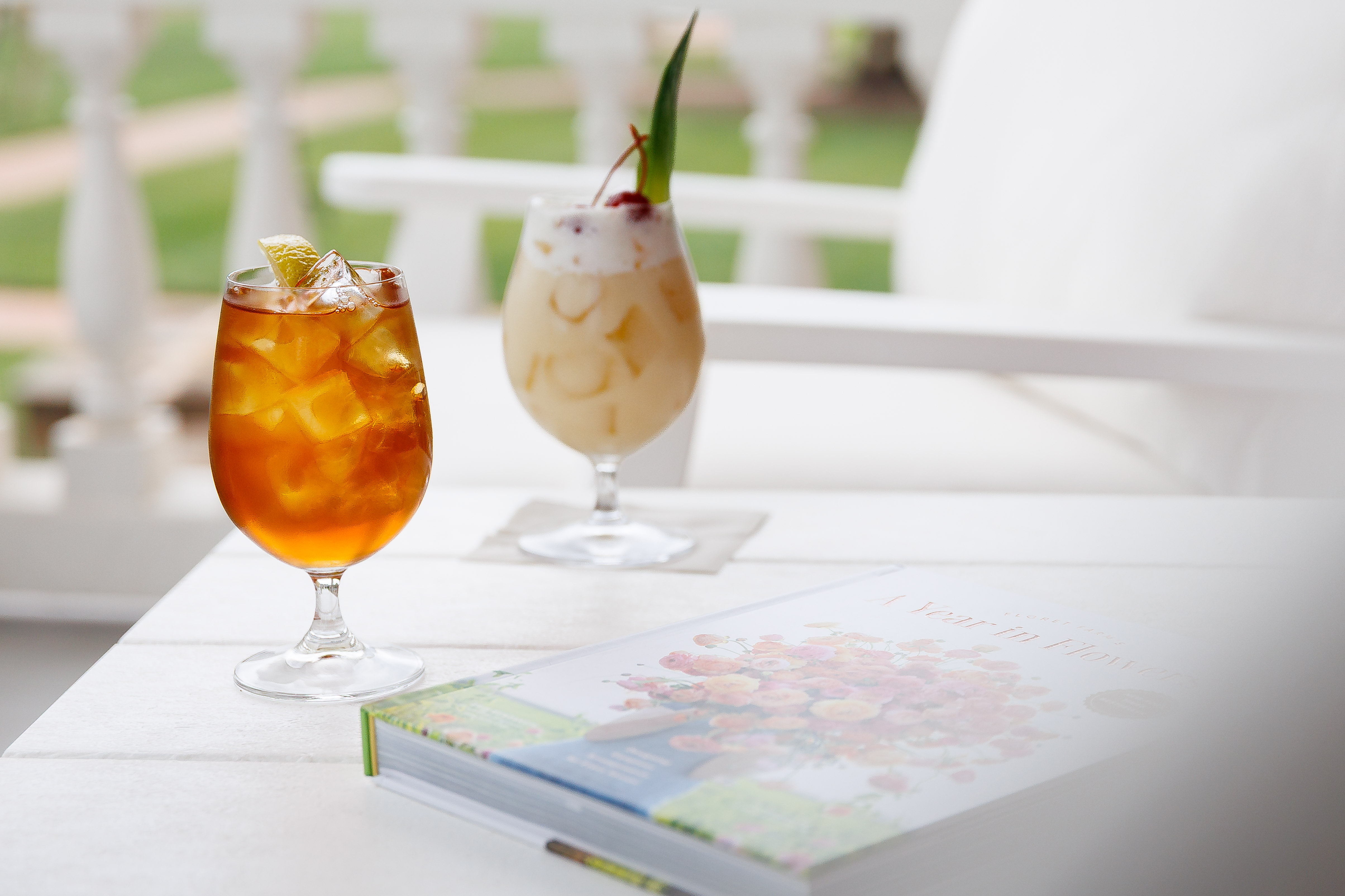 Two iced cocktails in tall glasses sit on a table, garnished with fruit, in a bright patio setting.
