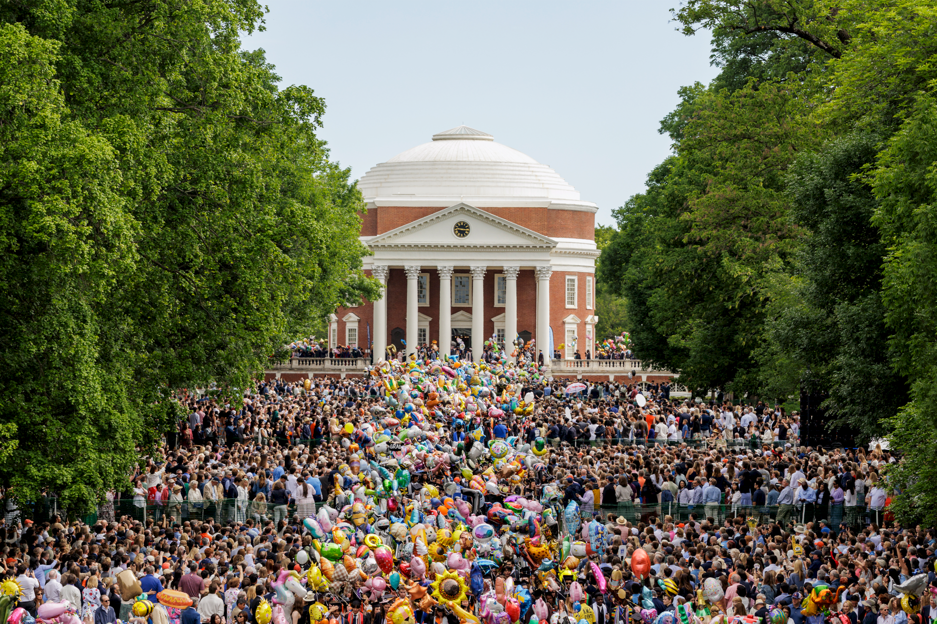 A huge crowd gathers in front of a brick, white-columned dome building as colorful balloons float over the celebration.